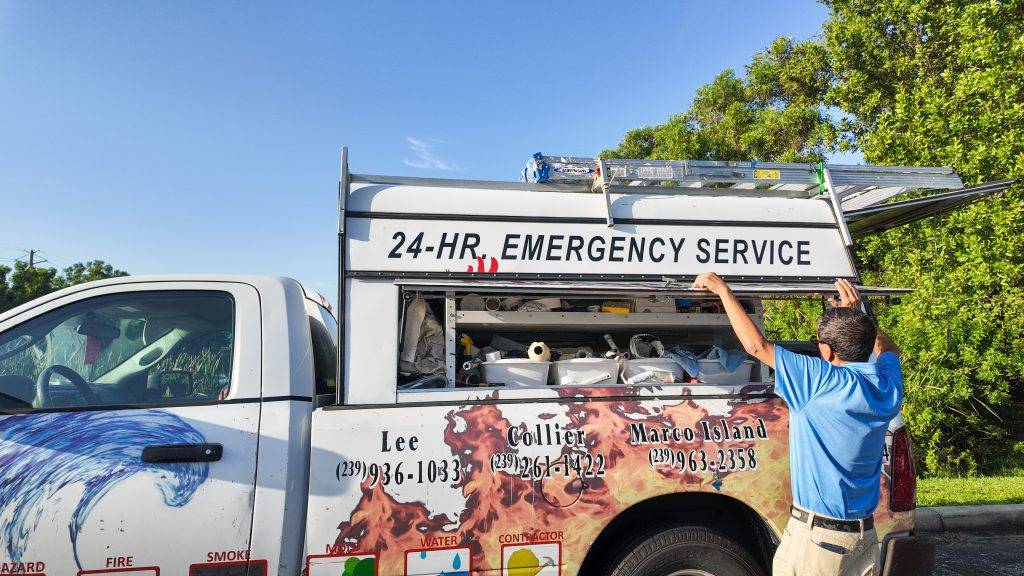 A person in a blue shirt opens the side compartment of a 24-hour emergency service truck, ready for storm clean-up, surrounded by trees.