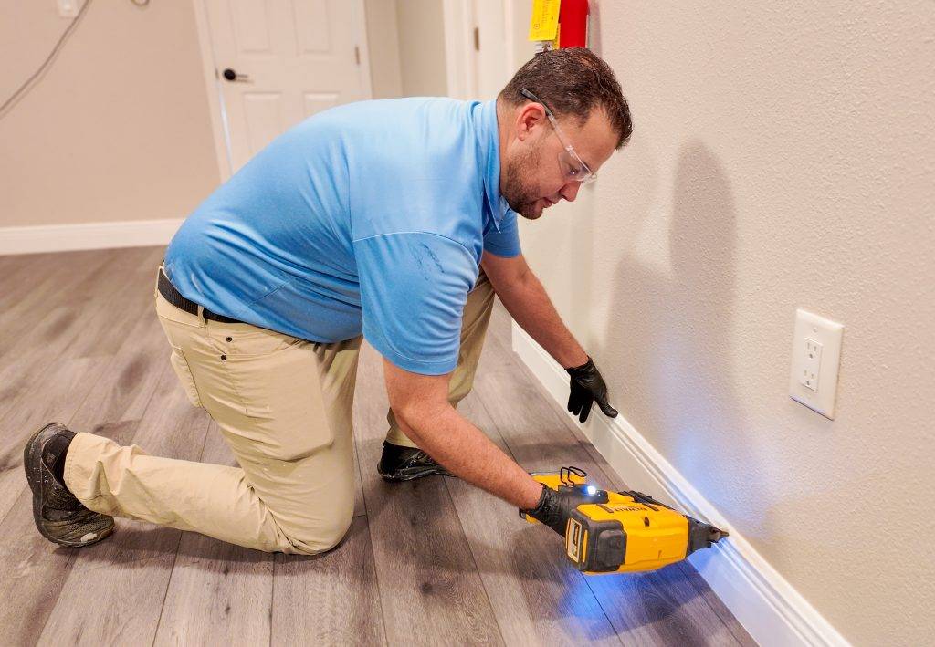 Wearing safety goggles, a man in charge of mold remediation installs baseboards on a gray wall using a yellow power nailer.