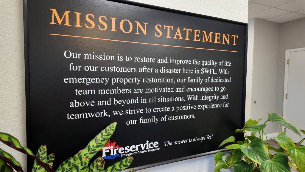 Framed mission statement sign on a wall, detailing a company's commitment to emergency fire and water damage restoration and customer service, with a small plant in the foreground.
