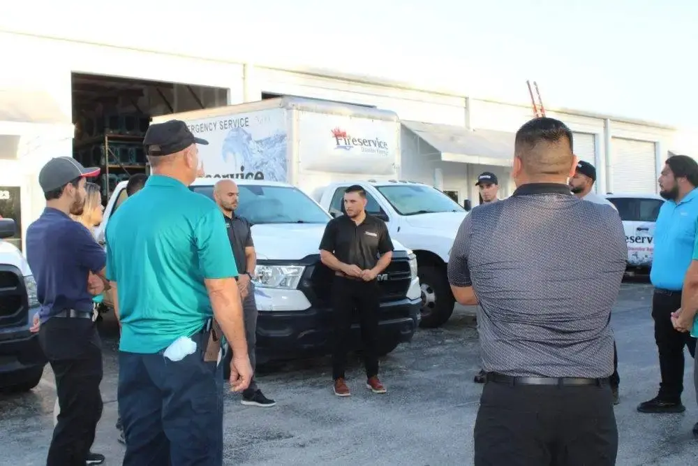 A group of people stands in a parking lot listening to a seated woman in front of several white vans.