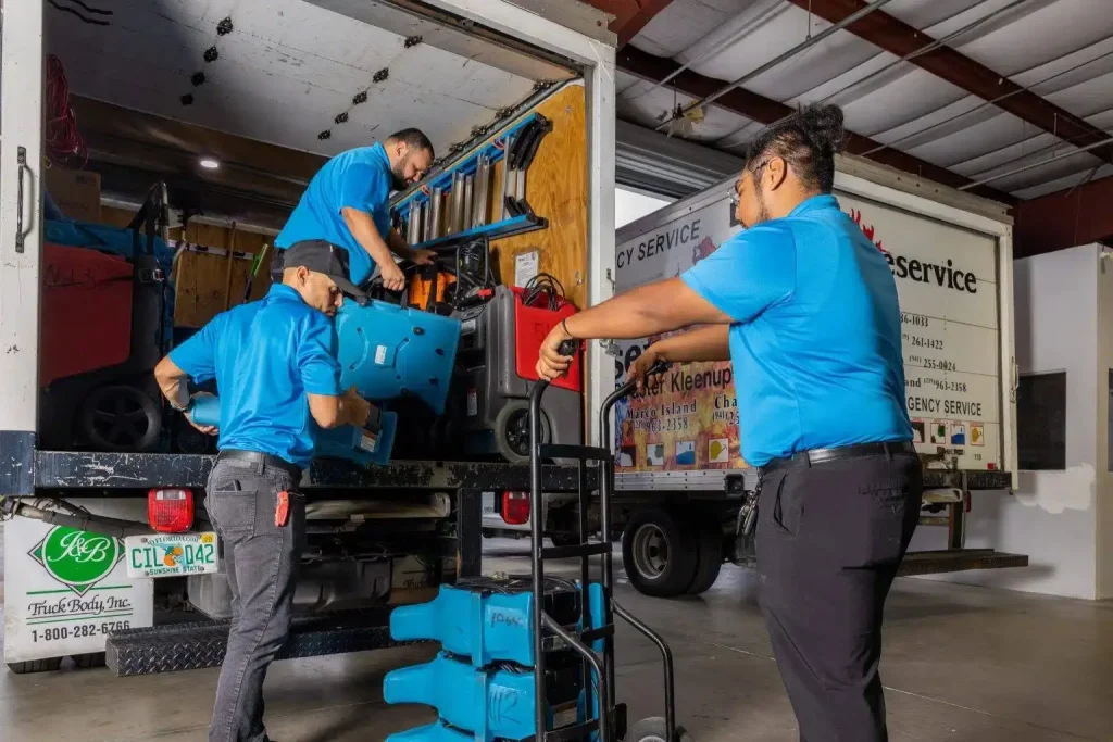 Three workers in blue shirts move blue equipment from a service truck inside a warehouse.