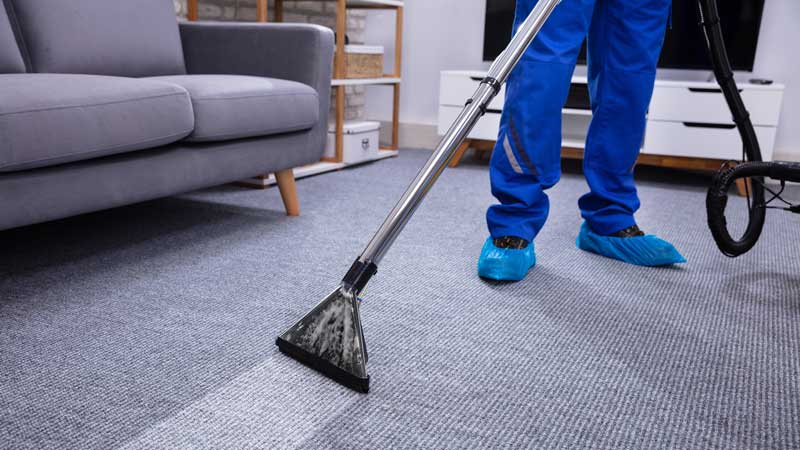 A person wearing blue scrubs and blue shoe covers cleans a gray carpet with a cleaning wand in a living room.