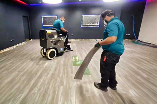 Two workers in teal shirts in a large indoor space; one holds a long curved black hose while the other stands near a cart with equipment.